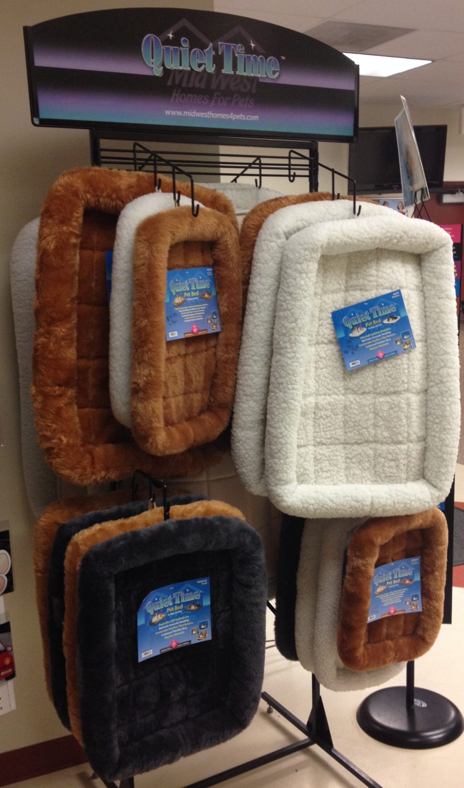 Four plush pet beds in various colors—brown, gray, and white—are displayed on a rack labeled "Quiet Time" in a pet store.